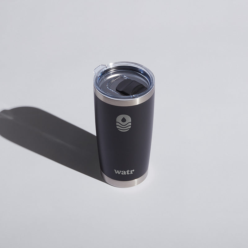 Alt text: Dark gray 20 oz WATR tumbler with a clear lid, featuring the WATR logo in white, positioned on a light gray surface with a distinct shadow to the right.