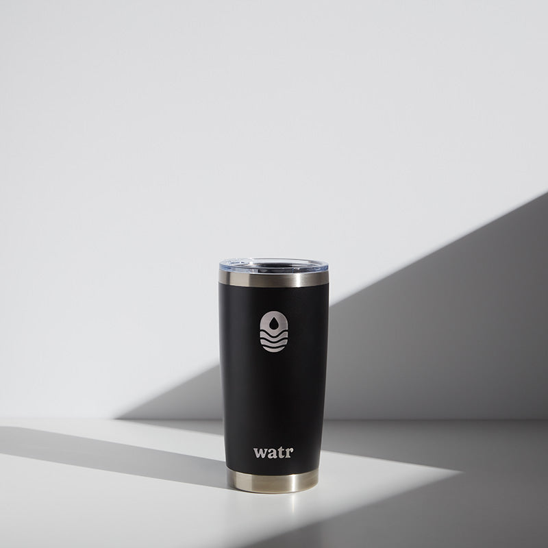 Alt text: "A 20 oz WATR Tumbler in black with a silver rim and clear lid, featuring a white minimalist wave logo above the brand name &