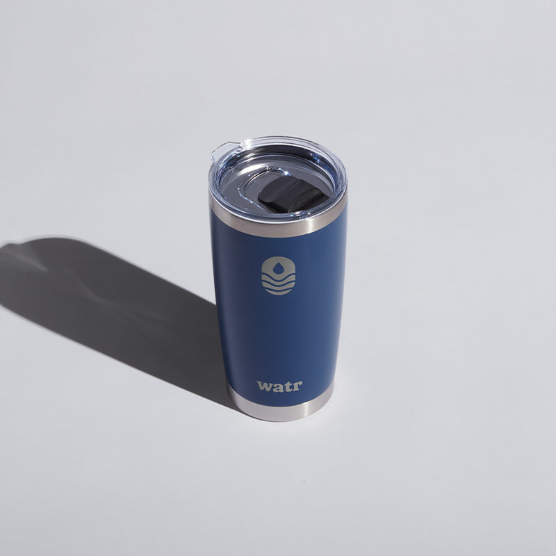 Blue 20 oz WATR tumbler with logo, featuring a clear lid, displayed on a light surface with a shadow to the side.