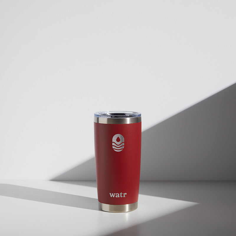 Red 20 oz WATR insulated tumbler with logo, stainless steel rim, and clear lid, presented in a softly lit environment with shadows.