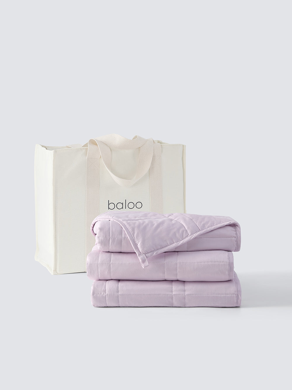 A folded weighted blanket in lavender color in front of a baloo tote bag on a white background [47867972059355]