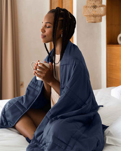 A young woman sits cross legged in a bed with a blue weighted blanket wrapped around her shoulders, and holds a warm cup of coffee. [43649942782171, 45574417875163, 47185819762907, 47185819992283, 47185820025051, 44422804340955, 44422811287771, 46301293543643, 46301293773019, 46301293805787, 46301293904091]