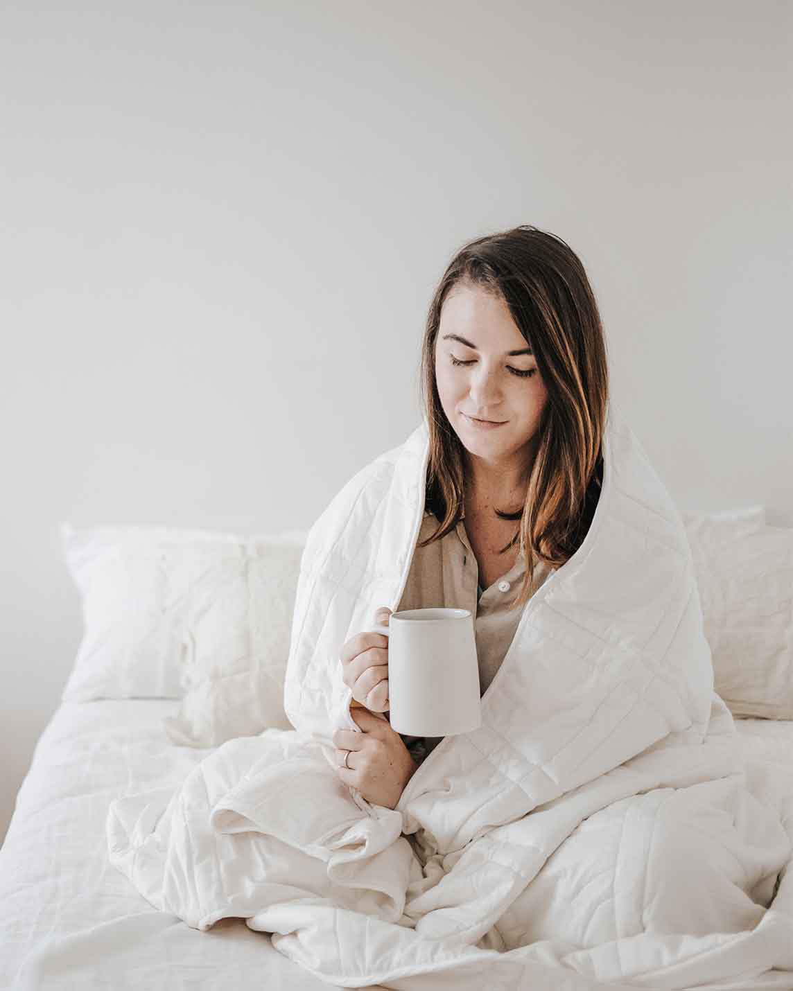 A young woman with brown hair sits cross legged on a bed with a white weighted blanket wrapped around her shoulders and holding a mug, softly smiling. [42934587621595, 45574417809627, 47185819697371]