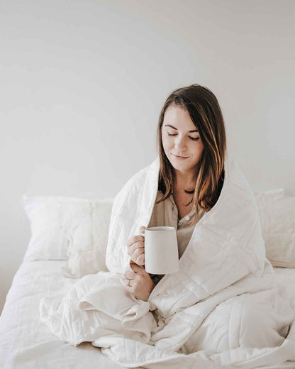 A young woman with brown hair sits cross legged on a bed with a white weighted blanket wrapped around her shoulders and holding a mug, softly smiling. [42934587621595, 45574417809627, 47185819697371]