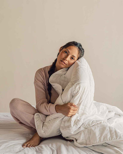 A young woman sits cross legged on a bed wearing soft pink pajamas and holding a white weighted blanket to her face. [42934587621595, 45574417809627,42934587883739, 46452819001563, 47185819697371, 47185819926747]