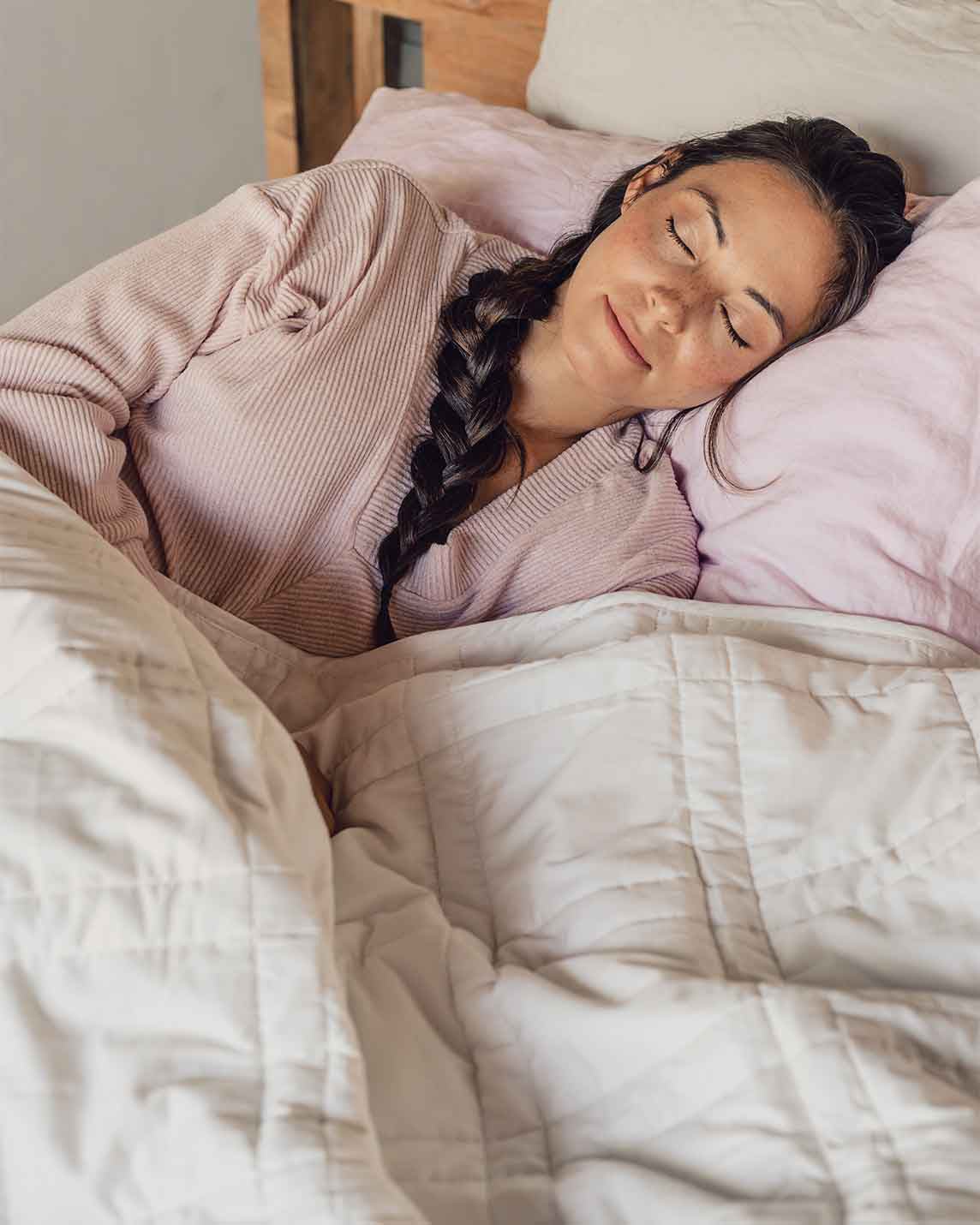 A young woman with a braid sleeps peacefully in a bed with a pink pillowcase and a white weighted blanket across her body.  [42934587621595, 45574417809627, 47185819697371, 46301293478107, 46301293576411, 46301293641947, 46301293707483]