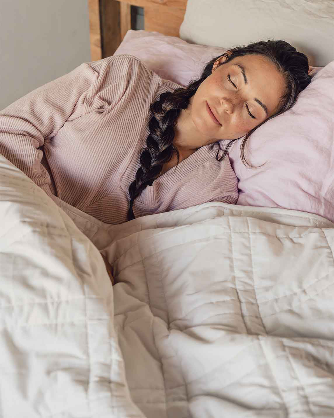 A young woman with a braid sleeps peacefully in a bed with a pink pillowcase and a white weighted blanket across her body.  [42934587621595, 45574417809627, 47185819697371]