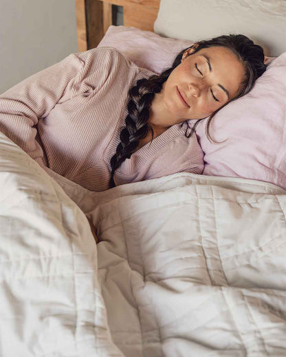 A young woman with a braid sleeps peacefully in a bed with a pink pillowcase and a white weighted blanket across her body.  [42934587621595, 45574417809627, 47185819697371]