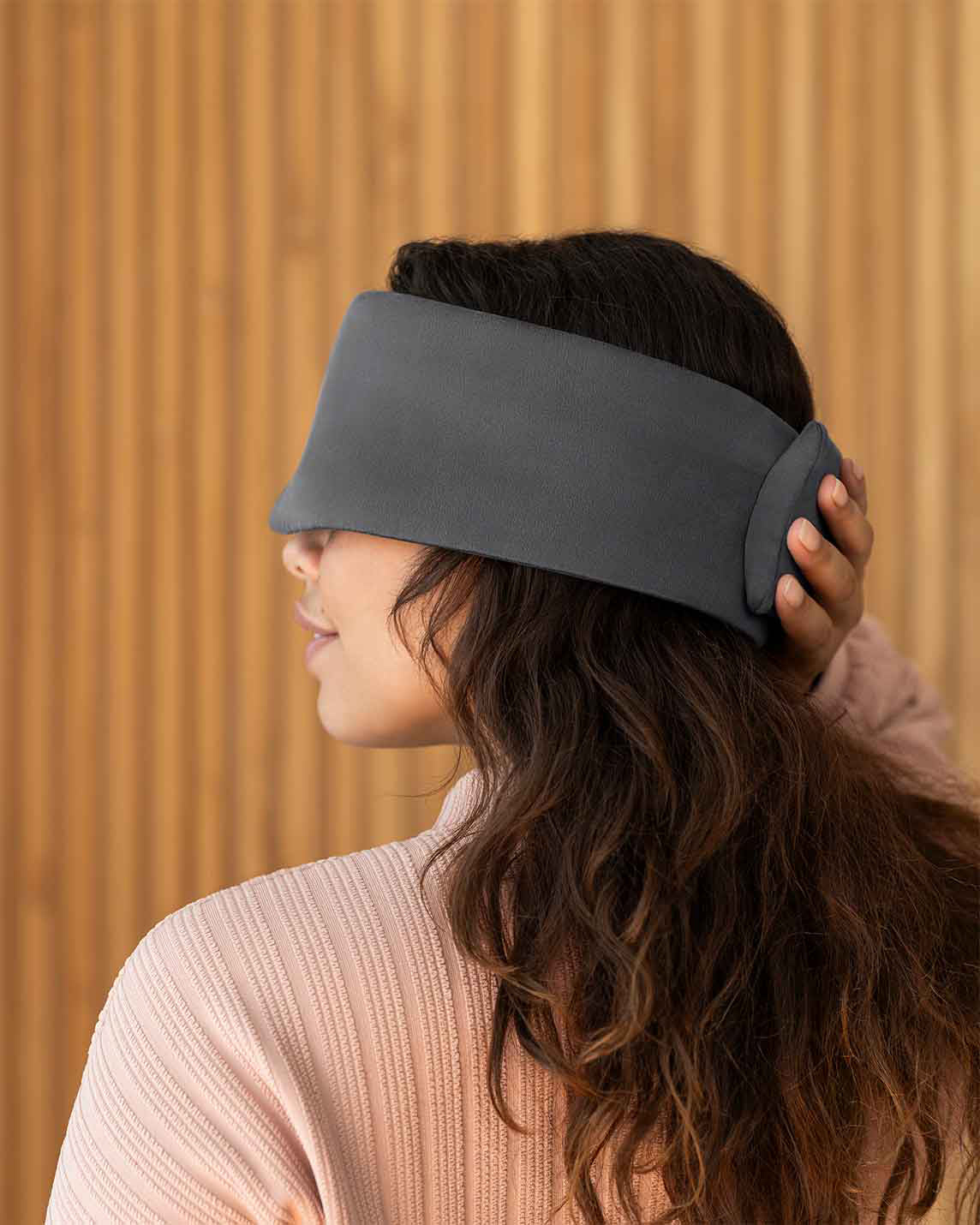A photo from the side of a woman with long brown hair holding her hand over the back of a silk sleep mask. [44377020793051, 47185819435227]