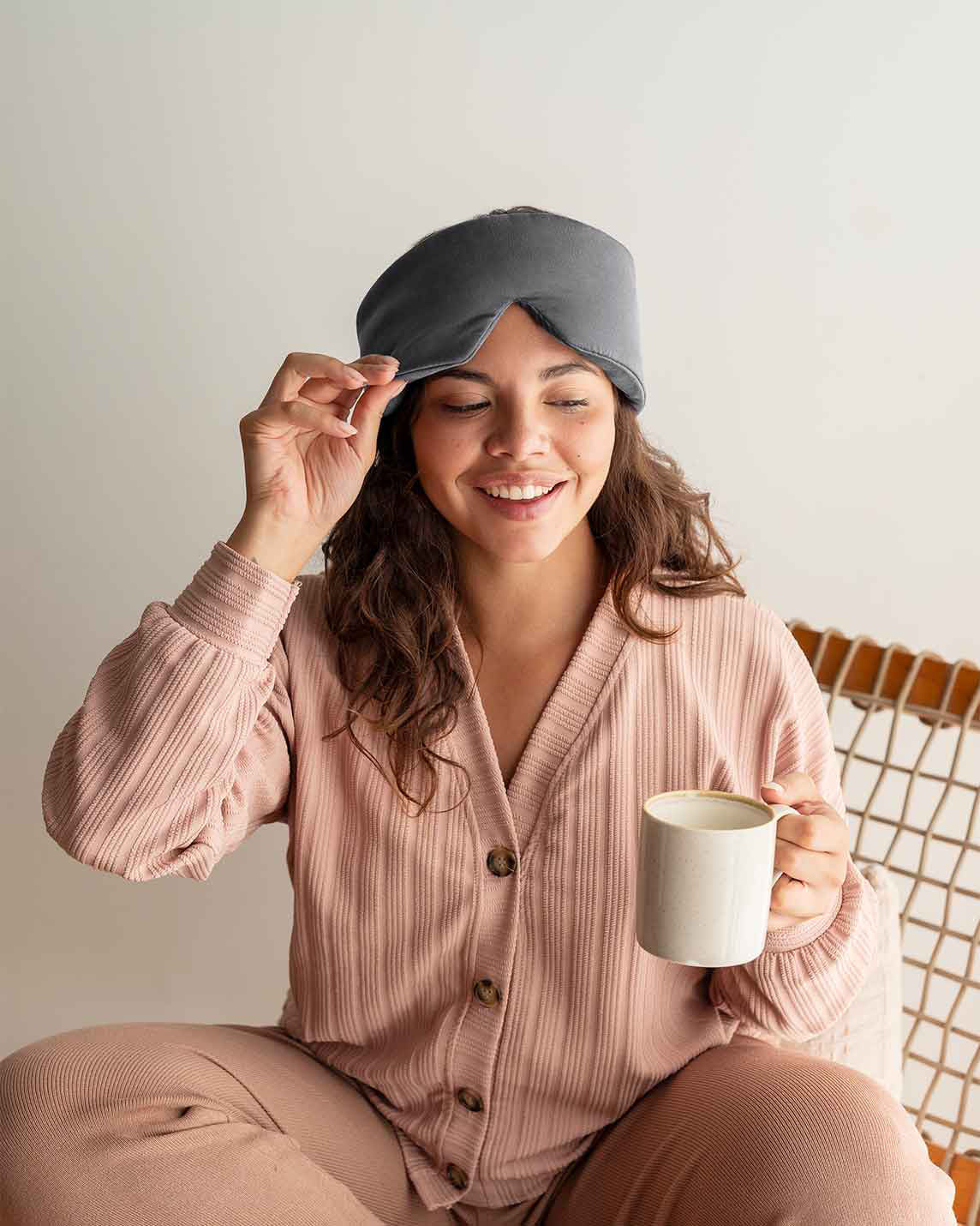 A woman with curly brown hair sits in a chair holding a mug, lifting the edge of a silk sleep mask up [44377020793051,46730820911323, 46730820976859, 46730821042395, 46730821107931, 47185819435227]
