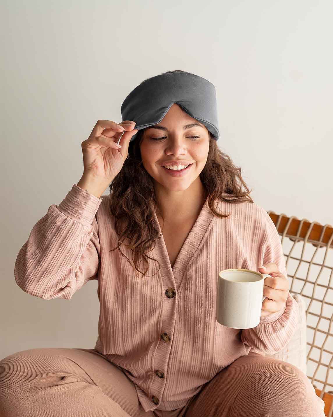 A woman with curly brown hair sits in a chair holding a mug, lifting the edge of a silk sleep mask up [44377020793051,46730820911323, 46730820976859, 46730821042395, 46730821107931, 47185819435227]