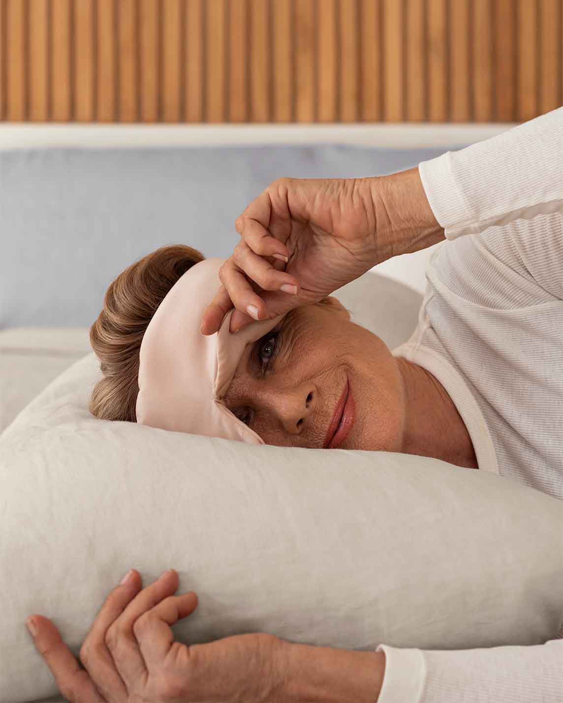 A senior woman with blonde hair lays down on a pillow wearing a pink silk sleep mask, holding one side up to view the camera [44377020825819, 47185819467995]
