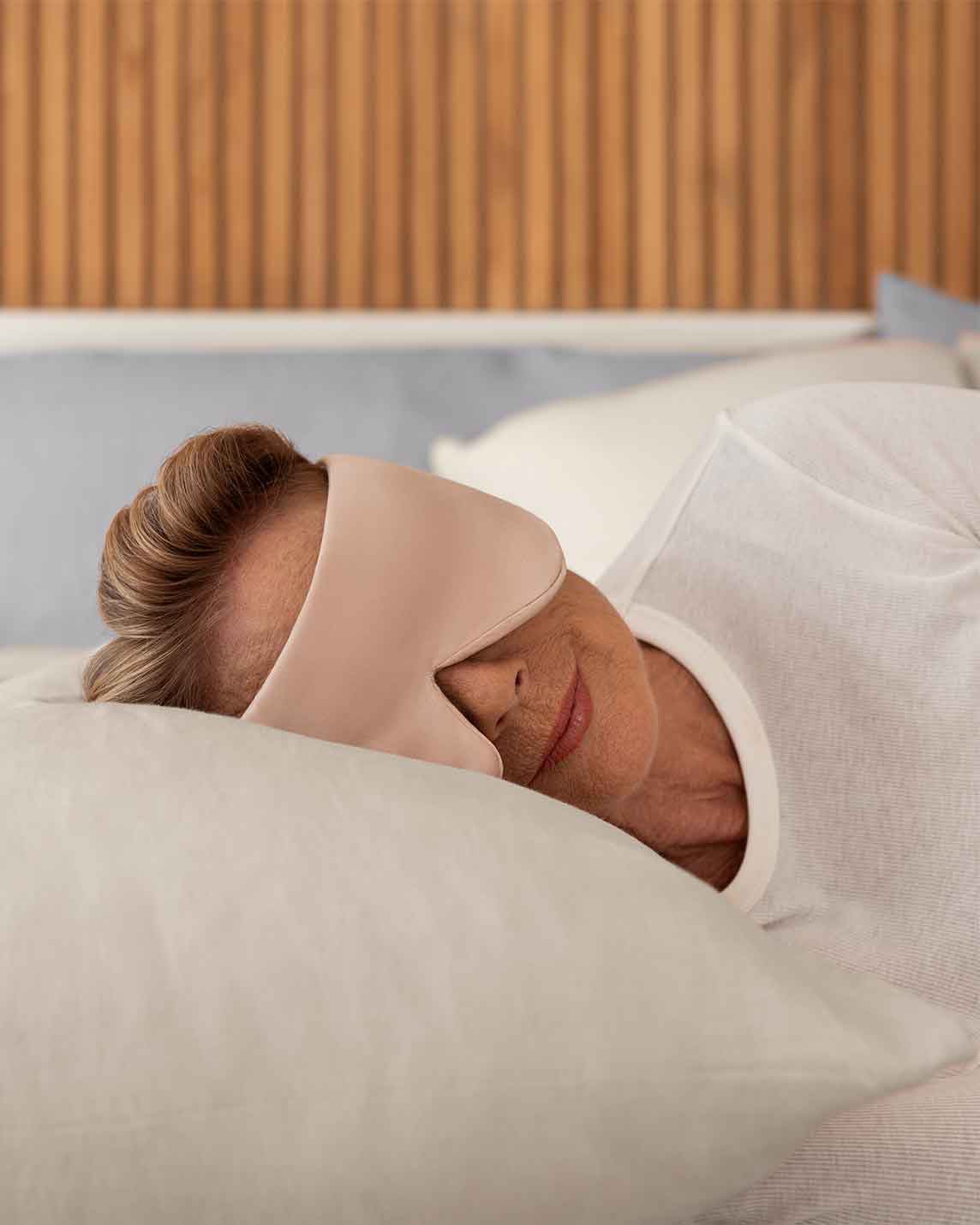 A senior woman with blonde hair rests her head on a pillow wearing a silk sleep mask [44377020825819,46730820944091, 46730821009627, 46730821075163, 46730821140699, 47185819467995]