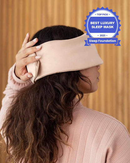 A photo from the side of a woman with long brown hair holding her hand over the back of a silk sleep mask. [44377020825819, 46730820944091, 46730821009627, 46730821075163, 46730821140699, 47185819467995]